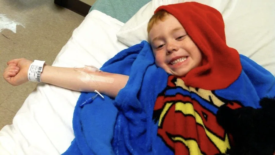 Child lying in hospital bed with IV line and Superman blanket during medical treatment.